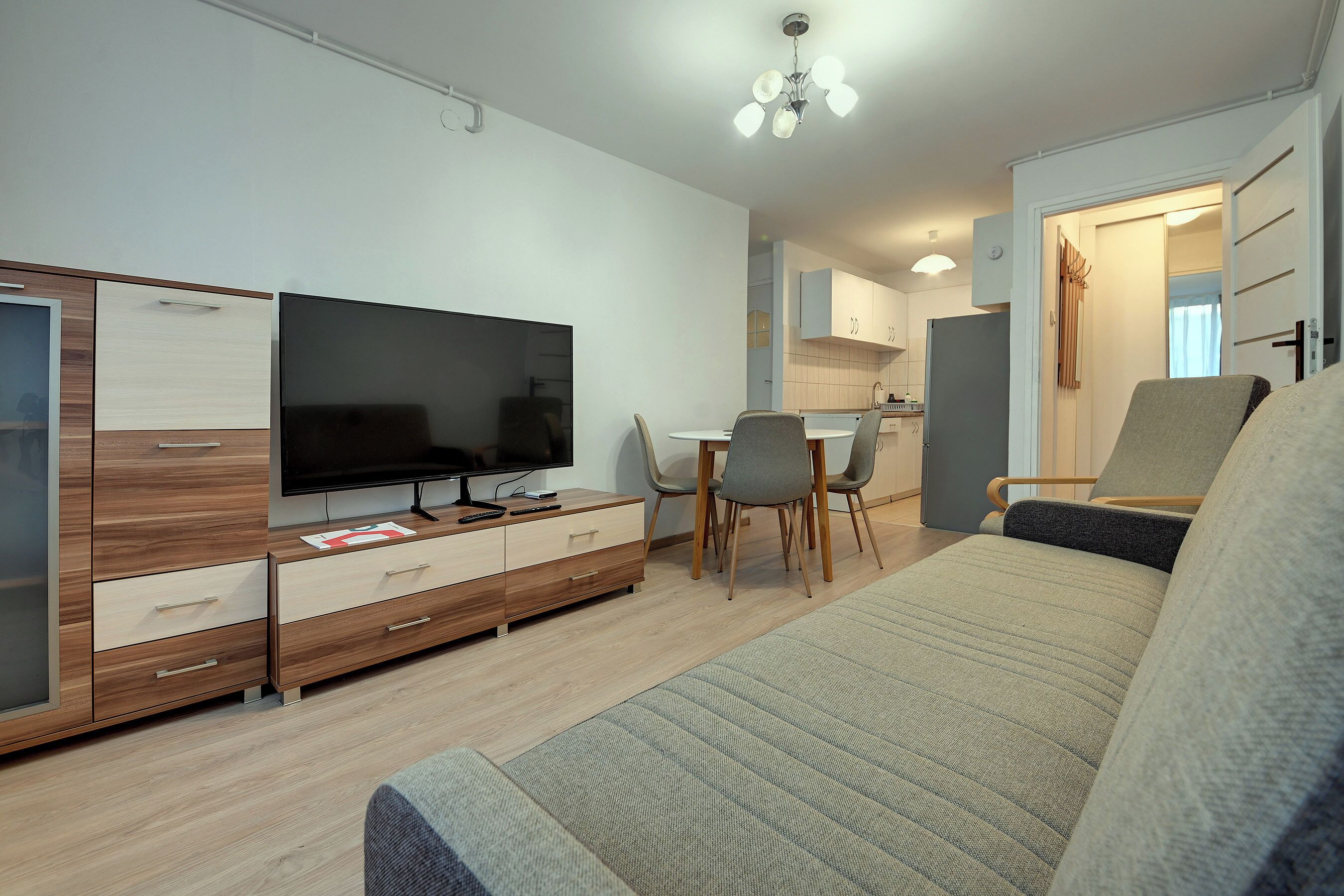 A modern living room with a flat-screen TV, wooden furniture, a dining area, and a kitchen in the background.
