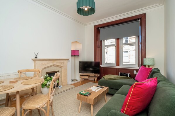 Een woonkamer met een open haard, een televisie, een groene bank en een eethoek met een tafel en stoelen.