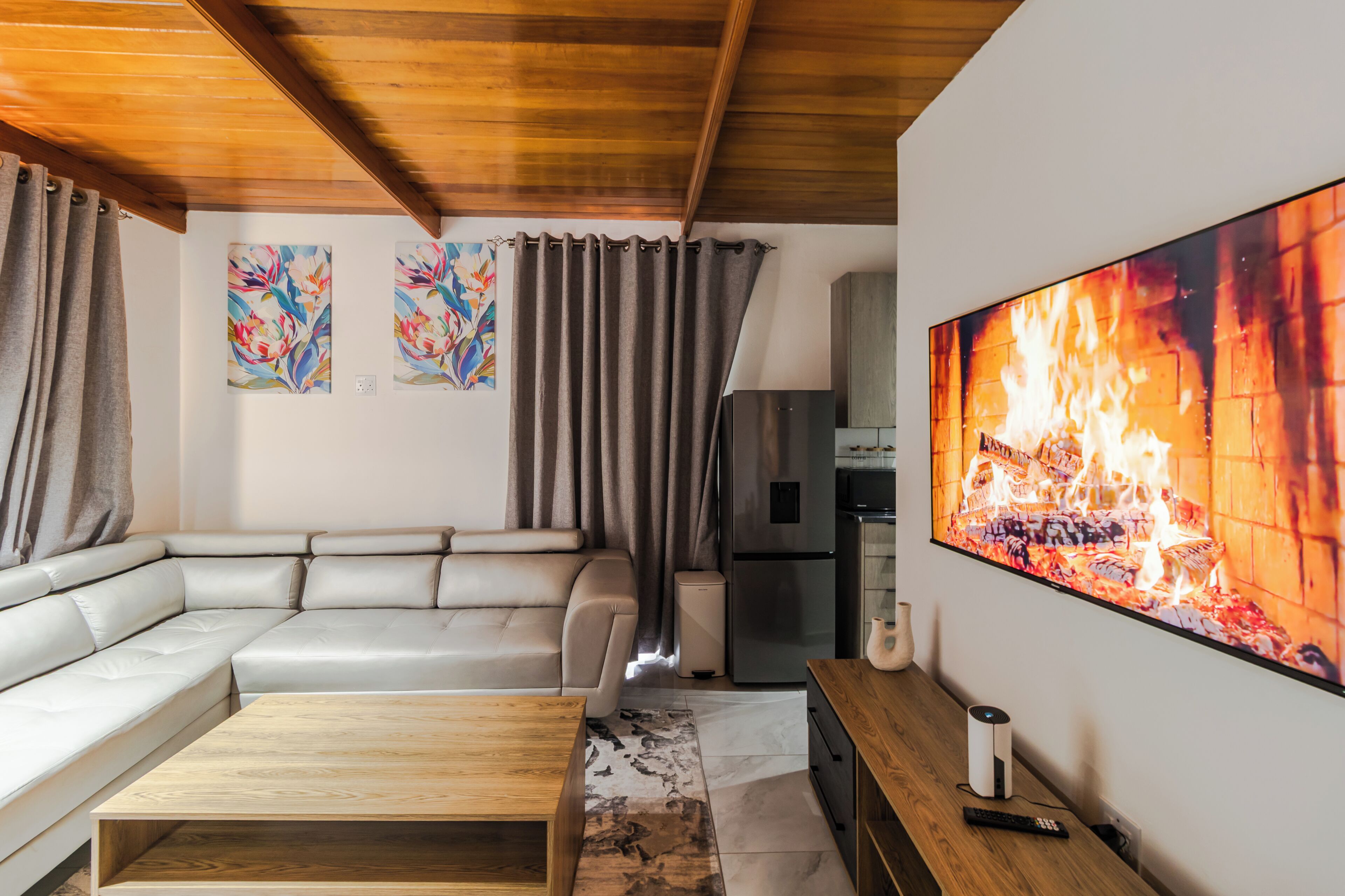 Uma sala de estar moderna com um sofá em formato de L, uma mesa de centro de madeira, uma televisão de tela plana exibindo uma lareira e uma área de cozinha com geladeira e micro-ondas.