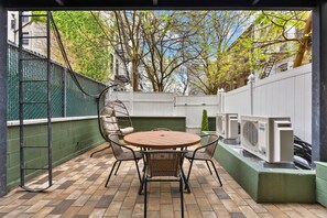 An outdoor patio with a round table and chairs, a green fence, and trees in the background. - Only in Brooklyn | 2BR + Backyard • Full Kitchen (Brooklyn)