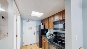 A compact kitchen with wooden cabinets, a black microwave, and a stainless steel refrigerator. - Camelot by the Sea: resort in beautiful Myrtle Beach. Fitness room, pool & beach (Myrtle Beach)