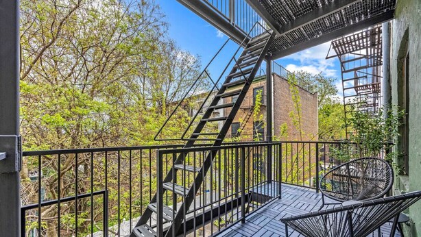 Ein Balkon mit Metalltreppe, einem Korbstuhl und Blick auf Bäume und Gebäude.