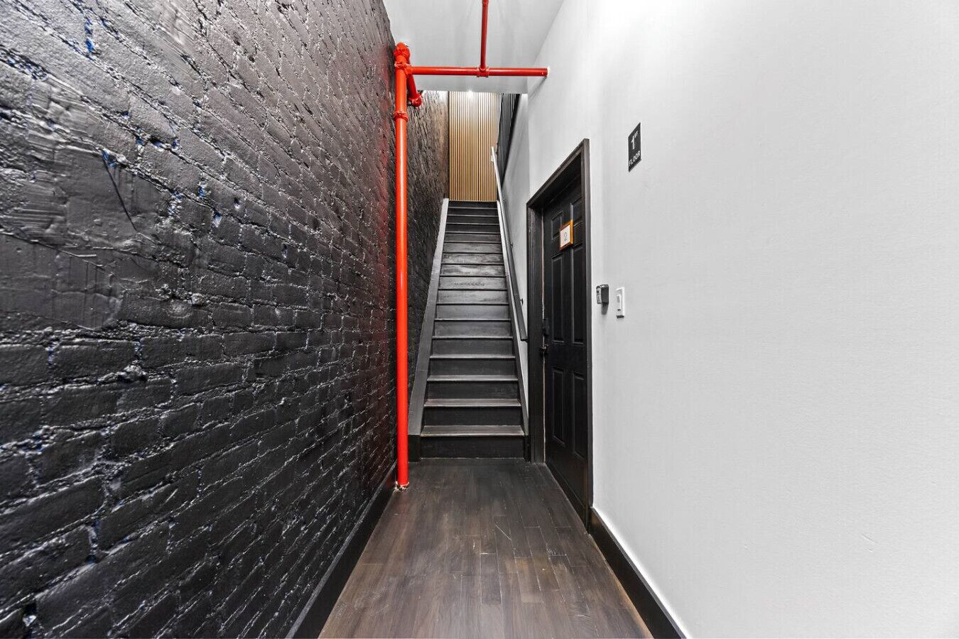 A narrow hallway with a black brick wall, a wooden floor, and a red pipe running along the wall.