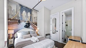A modern hotel room with a large bed, a cityscape mural, a wooden side table, and a walk-in closet.