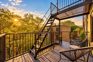 Ein Balkon mit Metalltreppe, einem Korbstuhl und Blick auf Bäume und Gebäude.
