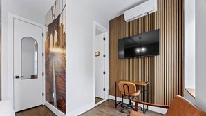 Ein modernes Wohnzimmer mit Flachbildfernseher an einer Wand aus Holzpaneelen, einem Schreibtisch mit Stuhl und einem großen Spiegel an der Tür.
