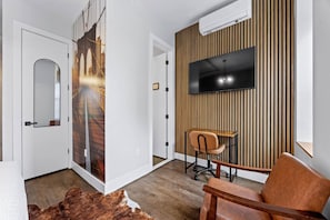 A modern living room with a flat-screen TV mounted on a wooden panel wall, a desk with a chair, and a large mirror on the door. - NYC 5 Borough Adventure | Family Stay + Balcony (Brooklyn)