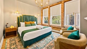 A hotel room with a large bed, a chandelier, a view of the outdoors through large windows, and a small seating area with a chair and a table.