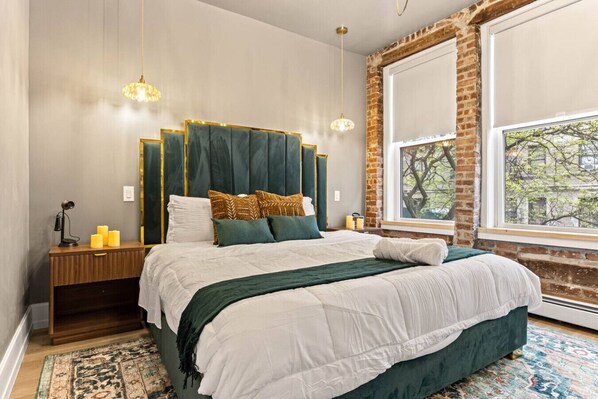 A bedroom with a large bed, a nightstand, a lamp, a window with a view, and a brick wall.
