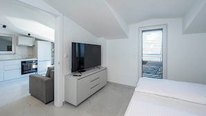 A modern bedroom with a large bed, a TV on a white console, a small seating area, and a kitchen in the background. - #3 Holiday Apartment MTG - Orebić (Orebić)