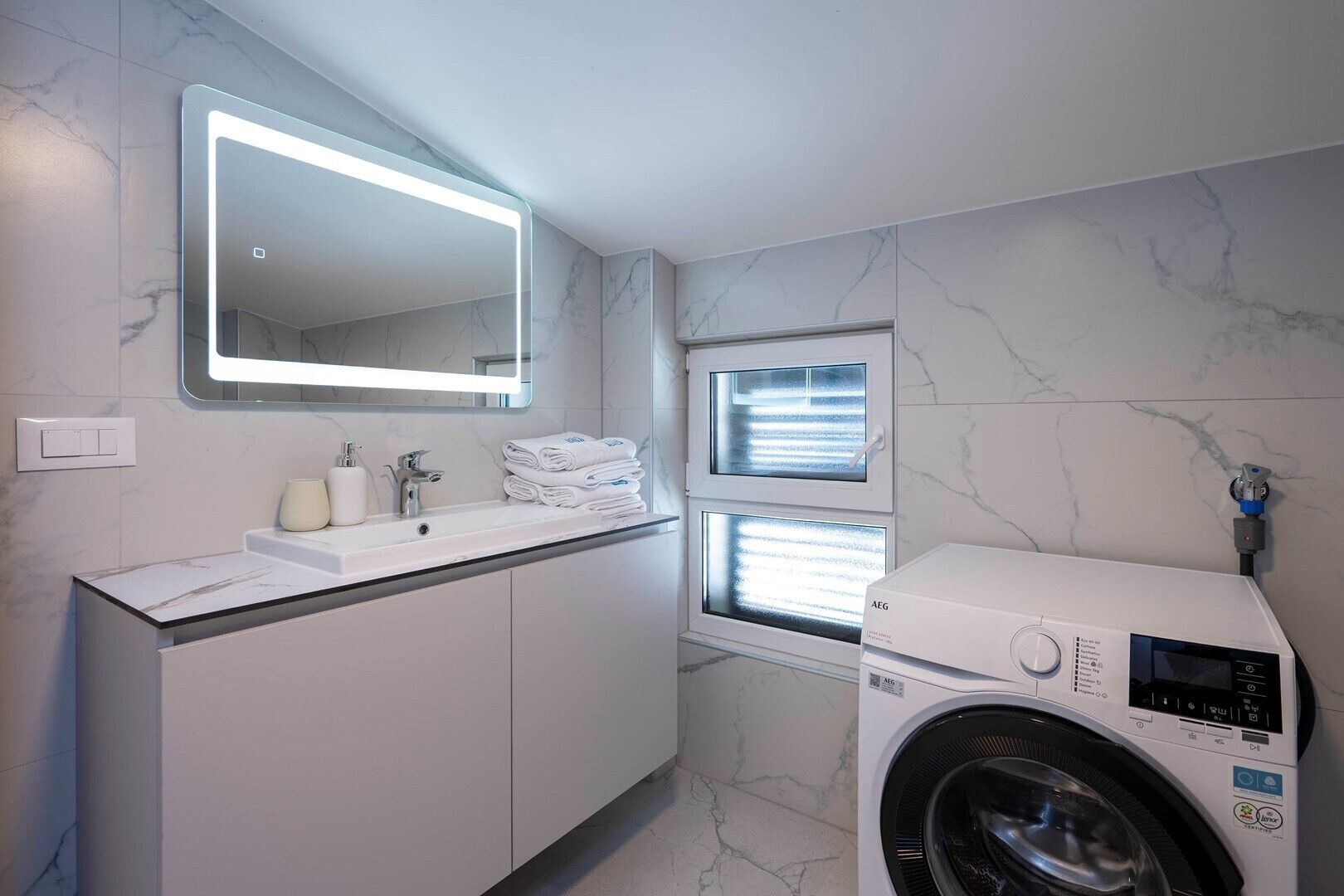 A modern bathroom with a built-in washing machine, a large mirror, and a window with blinds.