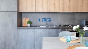 Moderni keittiö, jossa on harmaat kaapit, puinen työtaso, ruostumattomasta teräksestä valmistettu pesuallas, kaasuliesi ja yrityksen ’immobiliere STELLA’ logo.