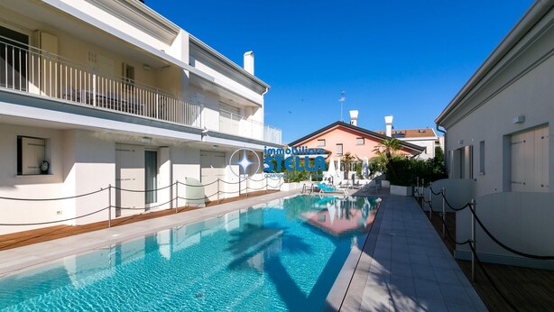 Une piscine avec une terrasse en bois, des bâtiments blancs et un ciel bleu azur.