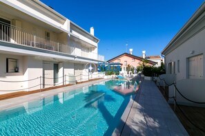 Une piscine avec une terrasse en bois, des bâtiments blancs et un ciel bleu azur.
