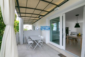 Eine überdachte Terrasse mit einem weißen Tisch und Stühlen, eine Glastür zu einem Innenbereich mit Essbereich, und das Logo von 'immobiliare STELLA'.