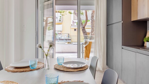 Een eetgedeelte met een tafel gedekt voor twee personen, een uitzicht door een glazen deur op een buitenbalkon, en een keuken met kasten en een aanrecht.