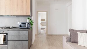 Een moderne keuken met een magnetron, fornuis en kasten, die uitkomt op een woonruimte met een bank en een plant.