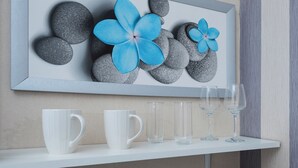 Una obra de arte en marco con flores azules sobre rocas, tazas blancas y vasos de vidrio sobre una estantería.