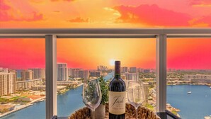 Une table avec une bouteille de vin et deux verres, placée sur un balcon donnant sur un paysage urbain et une étendue d’eau, au moment du coucher du soleil.