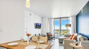 A modern living room with a dining area, a sofa, a TV mounted on the wall, and a large window with a view. - 2BR Resort Condo  High Floor Marina Views (Hallandale Beach)