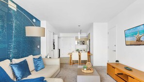 Ein modernes Wohnzimmer mit einer großen Weltkarte an der Wand, einer Couch mit blauen Kissen, einem Essbereich mit Tisch und Stühlen sowie einem Fernseher, der eine Strandansicht zeigt.
