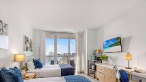 A hotel room with two beds, a desk with a computer, a TV, and a view of the city. - Sweeping Sunset Views -2BR Beachwalk Resort (Hallandale Beach)