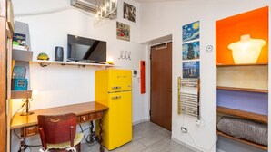 Una habitación pequeña con escritorio, un refrigerador amarillo, televisor y una puerta que da a otra habitación.