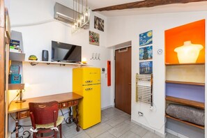 Ein kleiner Raum mit Schreibtisch, einem gelben Kühlschrank, einem Fernseher und einer Tür zu einem weiteren Zimmer.