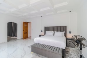 Ein modernes Schlafzimmer mit einem großen Bett, einem Nachttisch, einem Standventilator und einer Tür zu einem anderen Raum.