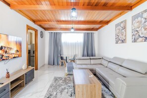 A modern living room with a wooden ceiling, a large flat-screen TV, a sectional sofa, a coffee table, and a dining area with chairs and a table. - Boutique Hotel-Style Room In Ferndale-G7 (Randburg)