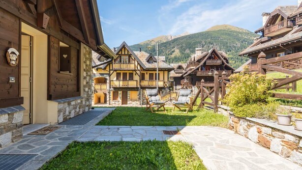 En traditionel chalet-bygning med en stenfacade, træbalkoner og en stikvej, der fører til et græsstykke med udendørs siddepladser.