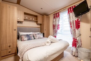 A bedroom with a bed, wooden wardrobe, built-in shelves, a television, and a window with curtains. - St Andrews Holiday Park | Harbour Reach (No 11) (St Andrews)