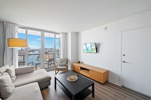 A modern hotel room with a sofa, a dining table, a chair, a TV on the wall, and a view of a cityscape through large windows. - Bright Apt, Intracoastal View | Balcony & Pool (Hallandale Beach)