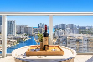 A table with a bottle of wine and two glasses on a tray, overlooking a cityscape with buildings and a body of water. - Intracoastal View Apt | Private Balcony & Full Kitchen (Hallandale Beach)
