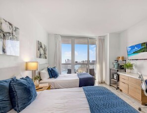 A modern hotel room with a bed, bedside table, lamp, and a view of the city through large windows. - Beachwalk 27th Fl | Private Bay Balcony (Hallandale Beach)