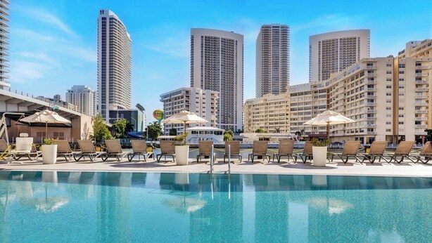 A hotel pool area with lounge chairs and umbrellas, surrounded by high-rise buildings. - 1BR · Intracoastal View · Balcony · Gym & Pool (Hallandale Beach)