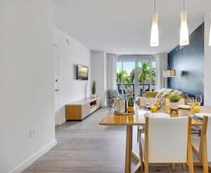 A modern dining area with a wooden table set for a meal, a TV mounted on the wall, and a view of the outdoors through large windows. - Above It All - 2BR - Intracoastal (Hallandale Beach)