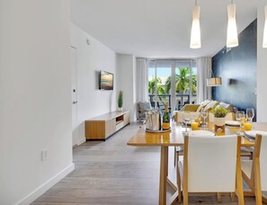 A modern dining area with a wooden table set for a meal, a TV mounted on the wall, and a view of the outdoors through large windows. - Sunset Views  2BR -2BTH (Hallandale Beach)
