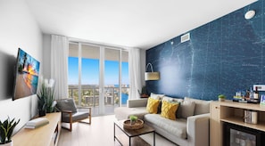 A modern living room with a large window, a flat-screen TV, a sofa, a coffee table, and a map wall. - Top-Floor 1BR · Bay View · Beachwalk Resort & Gym (Hallandale Beach)