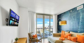 A modern living room with a TV, a sofa, a chair, and a view of the city through large windows. - Top-Floor 1BR · Bay View · Beachwalk Resort & Gym (Hallandale Beach)