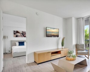 Ein modernes Wohnzimmer mit großem Fenster, einem Flachbildfernseher, einer Couch, einem Sessel und einem Holzschrank.