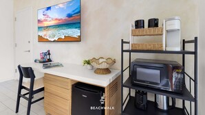 Ein Zimmer mit weißem Schreibtisch, einer Mikrowelle, einem Regal mit Kaffeemaschinen, einem Korbwannenstuhl und einem Bild einer Strandlandschaft.