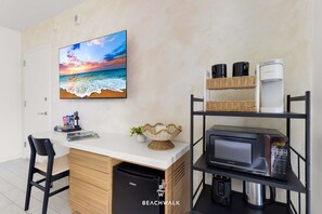 A room with a white desk, a microwave, a shelf with coffee makers, a wicker basket, and a painting of a beach scene. - Resort Living  2BR - Intracoastal View (Hallandale Beach)