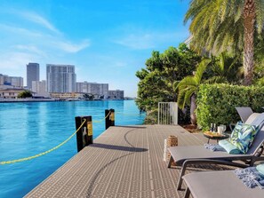 A waterfront balcony with lounge chairs, a pool, and a city skyline in the background. - Upper-Floor Bay View · 2BR Beachwalk Resort (Hallandale Beach)
