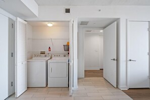 A laundry room with two washing machines and a dryer, a white door, and a hallway leading to another room. - Upper-Floor Bay View · 2BR Beachwalk Resort (Hallandale Beach)