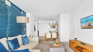 A modern living room with a large map on the wall, a sofa with blue cushions, a dining area with a table and chairs, and a kitchen in the background. - Stylish High-Floor 2BR · Beachwalk Resort (Hallandale Beach)
