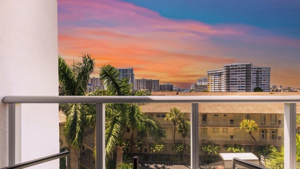 Un balcon offrant une vue sur le paysage urbain au coucher du soleil, avec des bâtiments modernes et des palmiers.