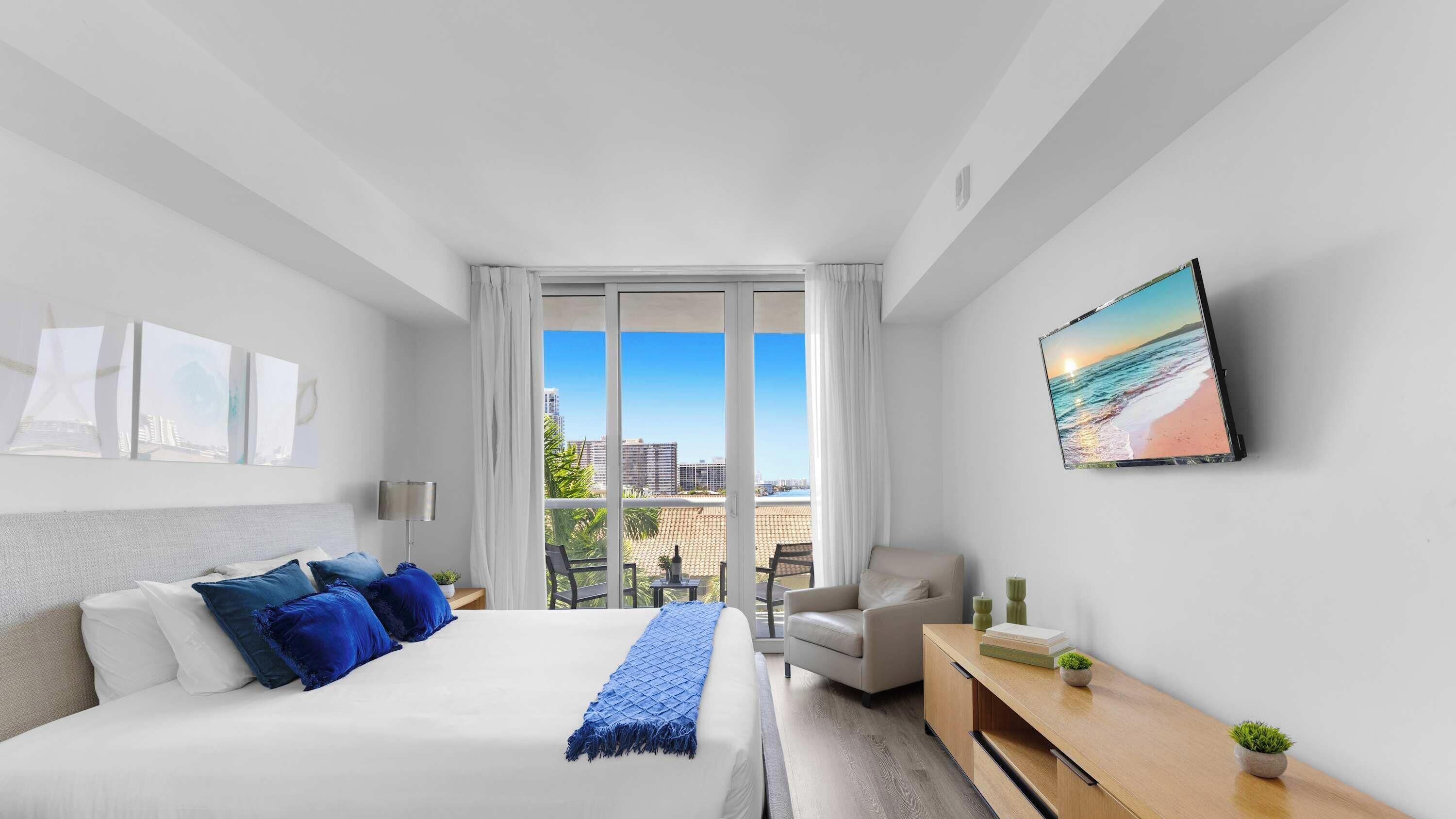 A hotel room with a bed, a chair, a desk, and a large window offering a city view.