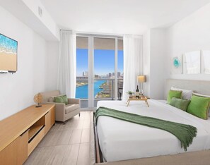 A modern hotel room with a large bed, a desk, a chair, and a view of a cityscape through large windows. - 1BR Balcony · Bay View · Beachwalk Resort (Hallandale Beach)
