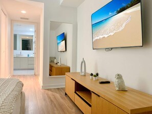 Een moderne woonkamer met een grote flatscreen televisie aan de wand, een houten console tafel met decoratieve objecten, en uitzicht op een strand door het raam.